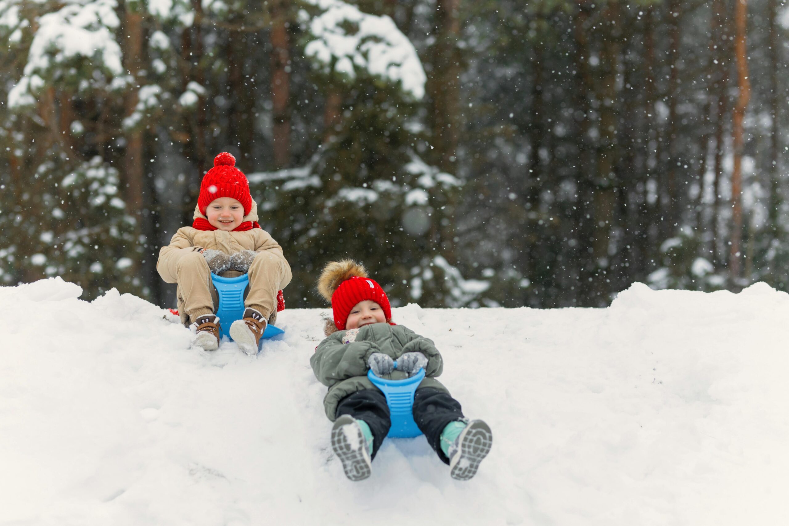 Two happy children wearing red hats sledding down a snowy hill in a winter forest.