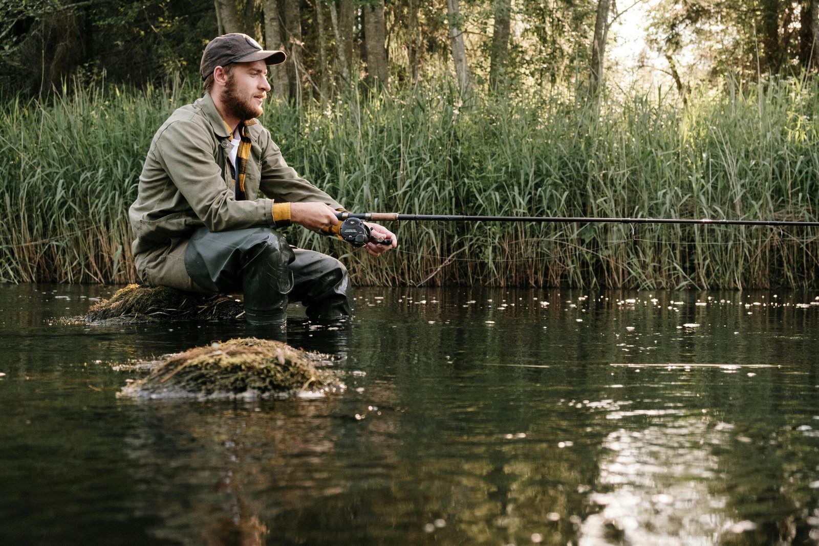 A man sitting peacefully by a stream fishing among reeds, reflecting on a calm summer's day.