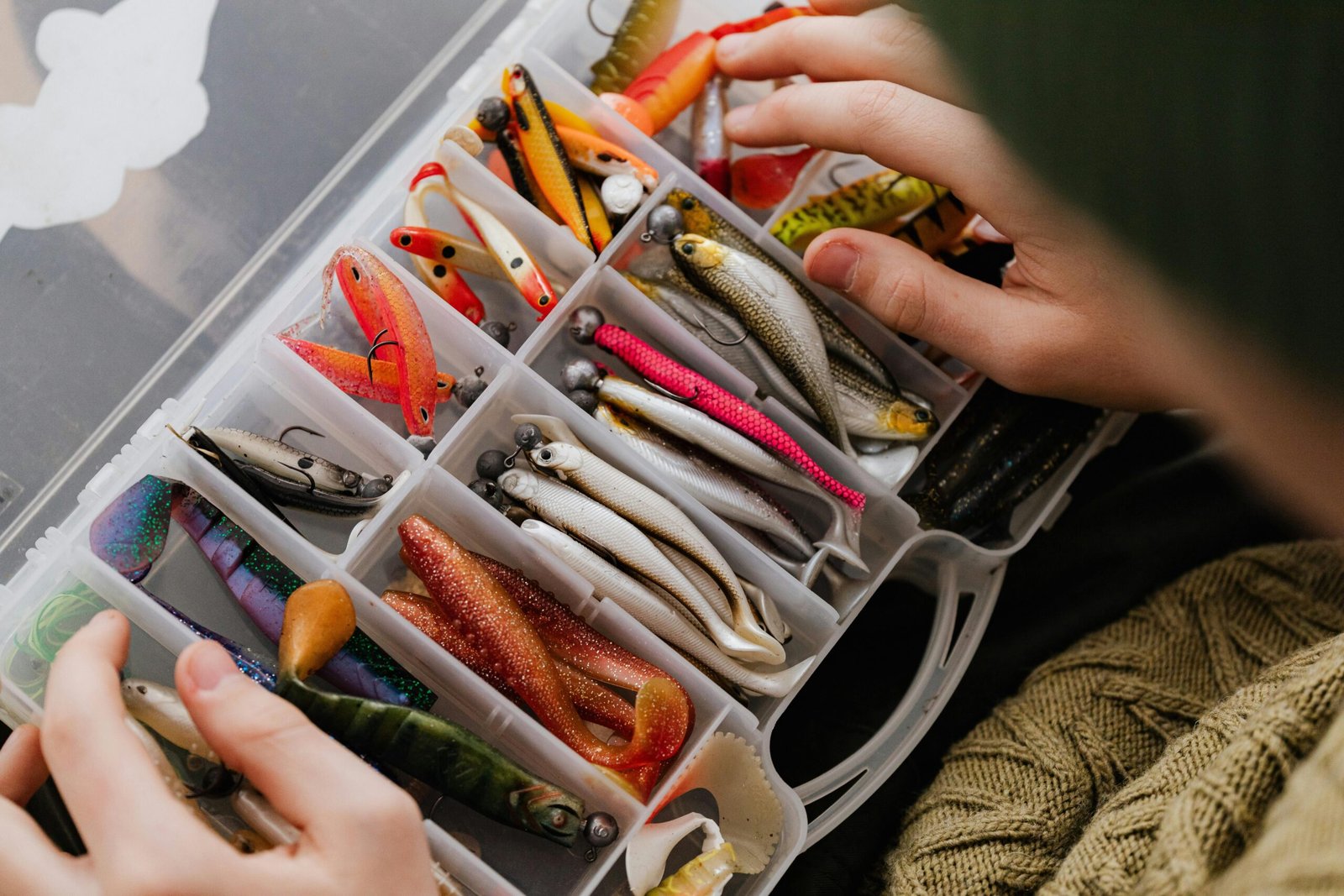 Hands organizing assorted fishing lures in a plastic box.